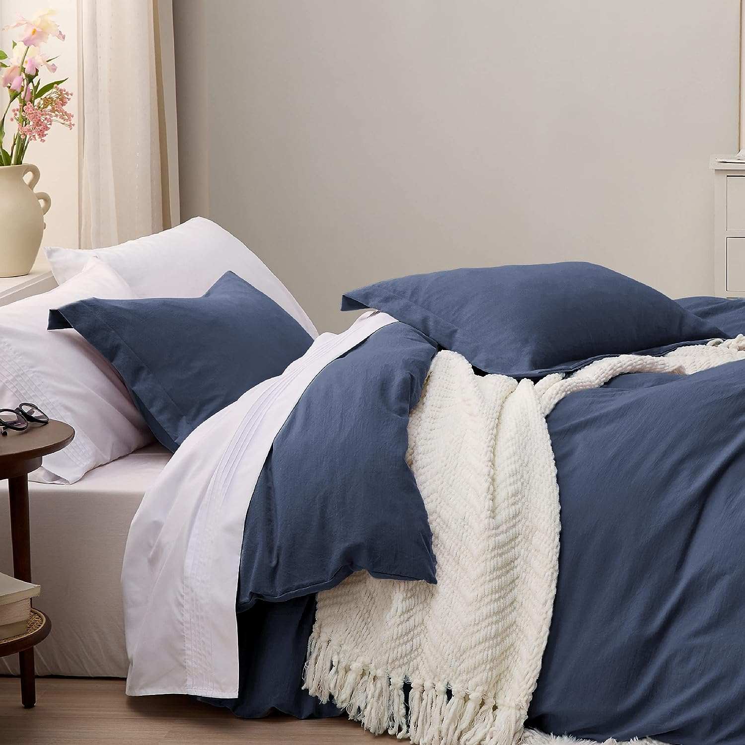 Cozy bedroom with navy blue bedding, white sheets, and a fringed blanket on a neatly made bed next to a small wooden side table with flowers.