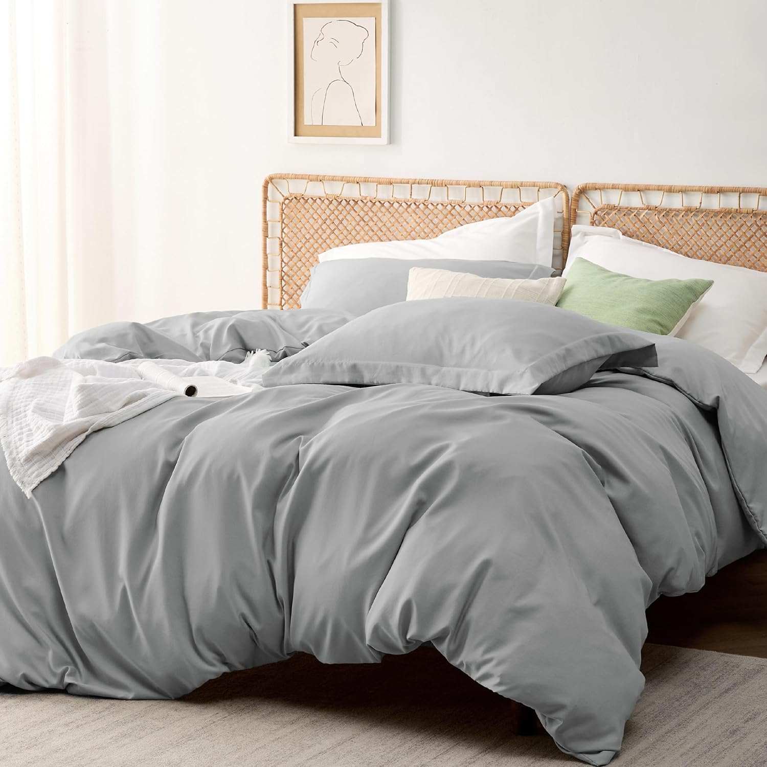 Cozy bedroom with a neatly made bed featuring gray bedding, wicker headboard, and decorative pillows. Minimalist wall art and soft natural lighting.