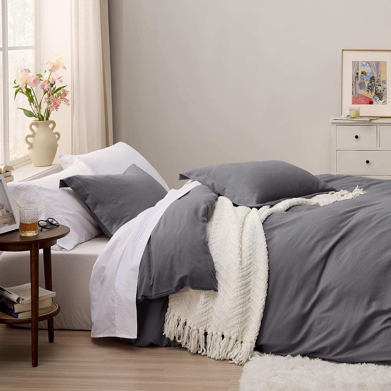 Cozy bedroom with grey bedding, white throw blanket, and a side table with flowers in a vase next to a large window with sunlight.