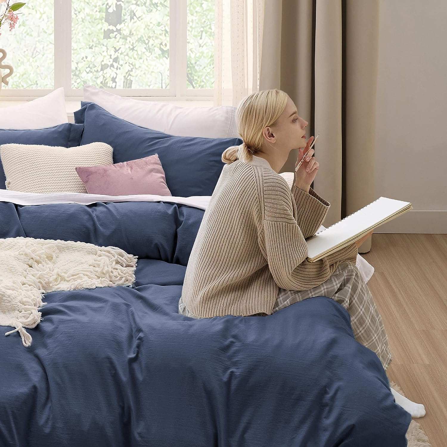 Woman sketching in a cozy bedroom with blue and white bedding, near a window with a serene view.