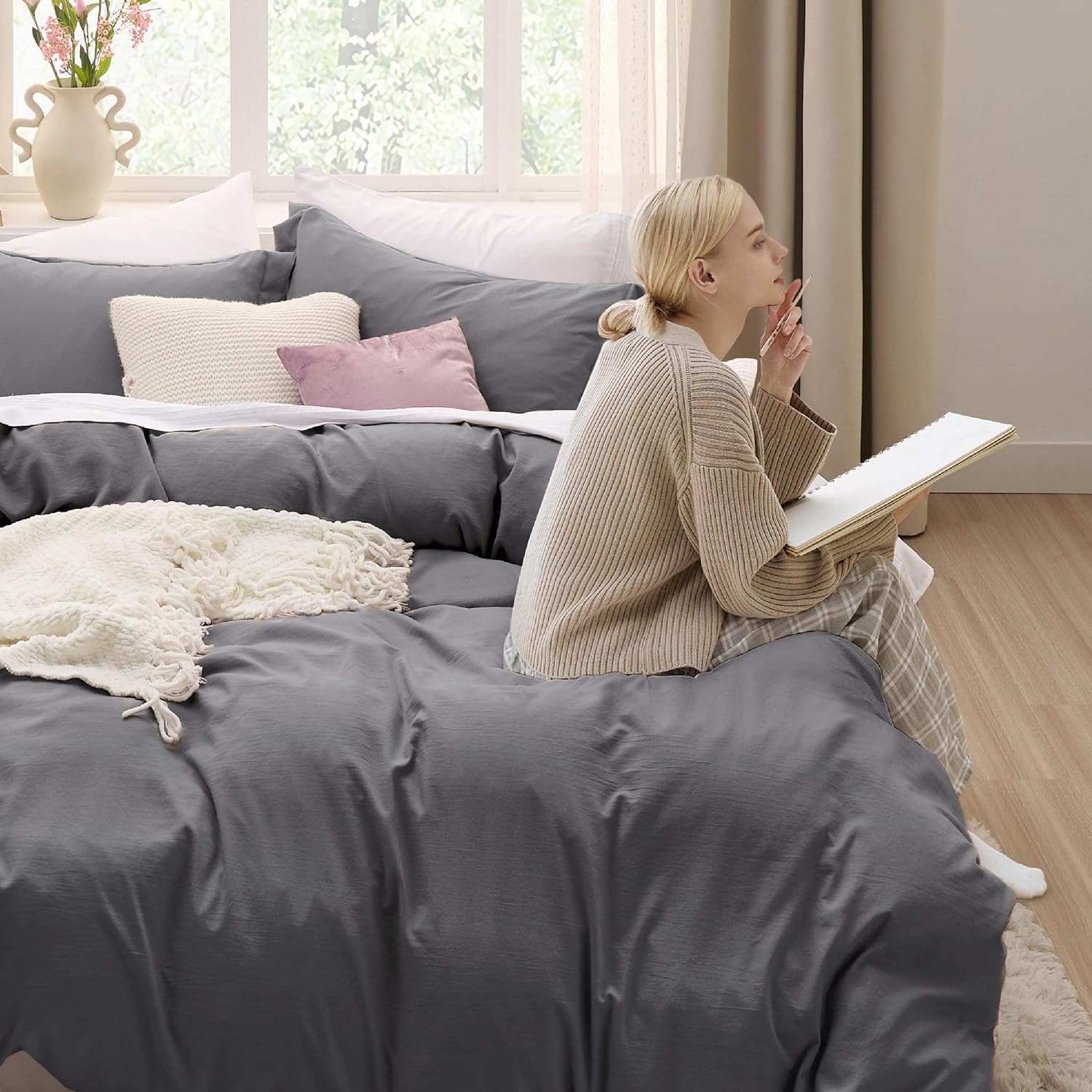 Woman sitting on a cozy bed with gray bedding, reading a book near a bright window with plants in the background.