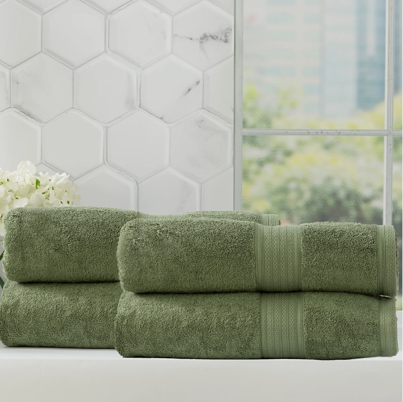 Set of green bath towels stacked in a modern bathroom with white hexagonal tiles and a window view of greenery.