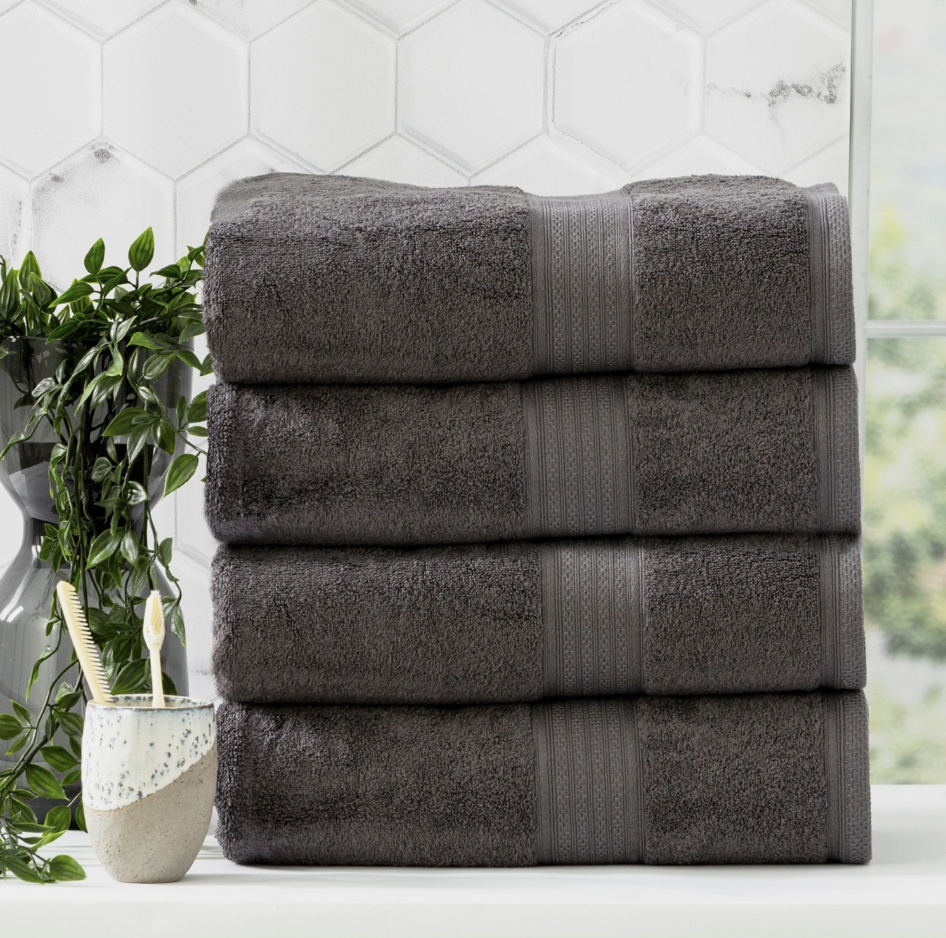 Stack of four plush dark gray towels with a potted plant and toothbrush holder next to a hexagonal tiled wall background.