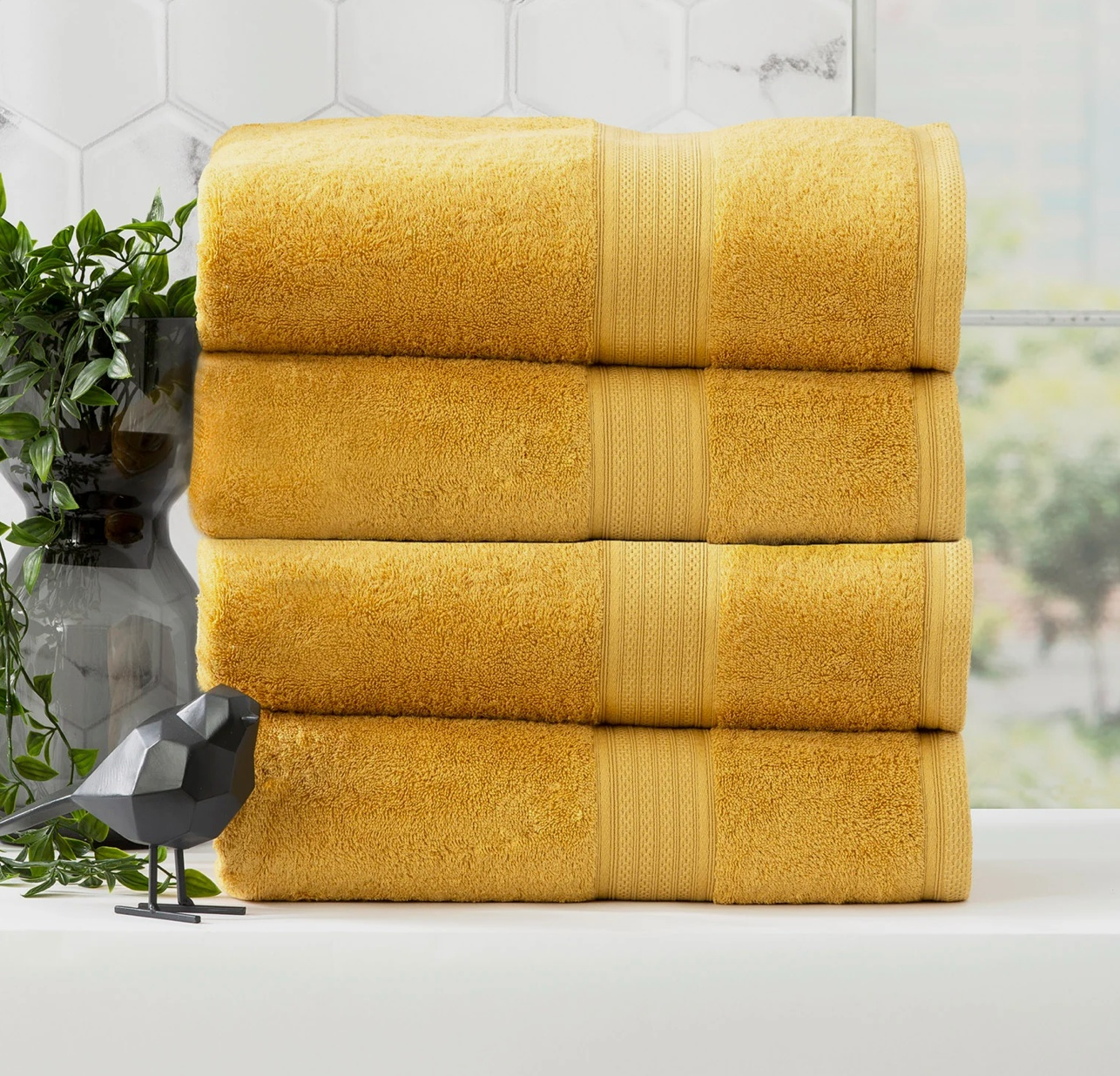 Stack of four vibrant yellow bath towels with a decorative black bird and green plant in the background in a bright bathroom.