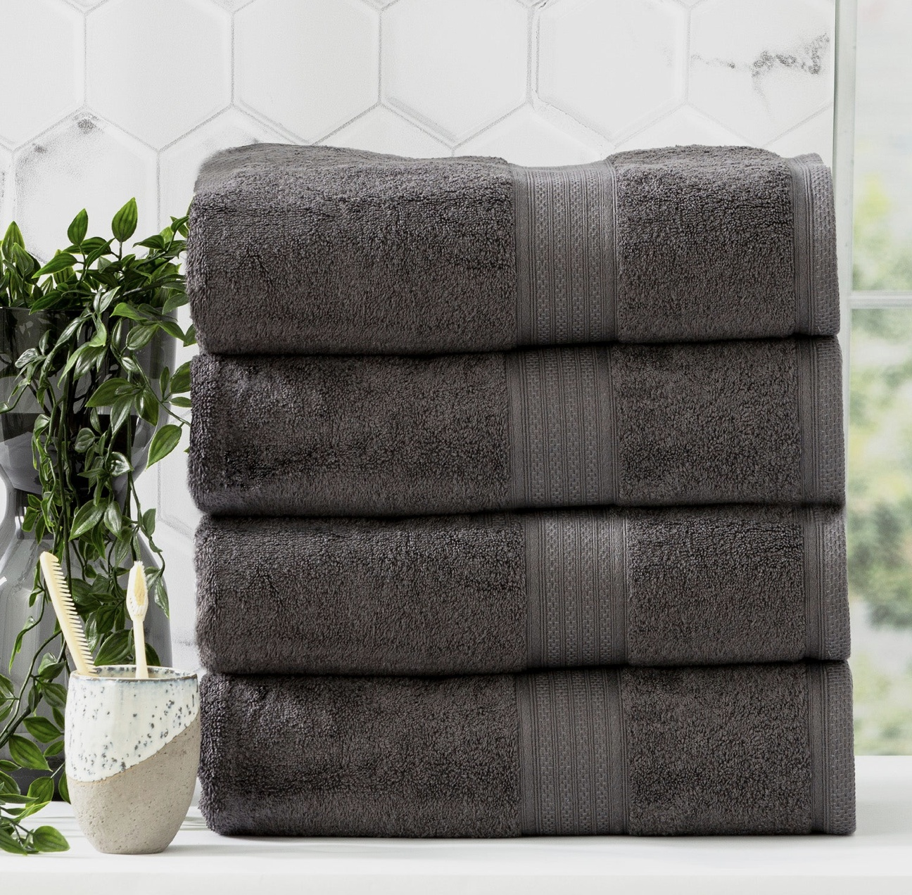 Stack of four plush gray towels on a bathroom counter with a potted plant and a toothbrush holder nearby