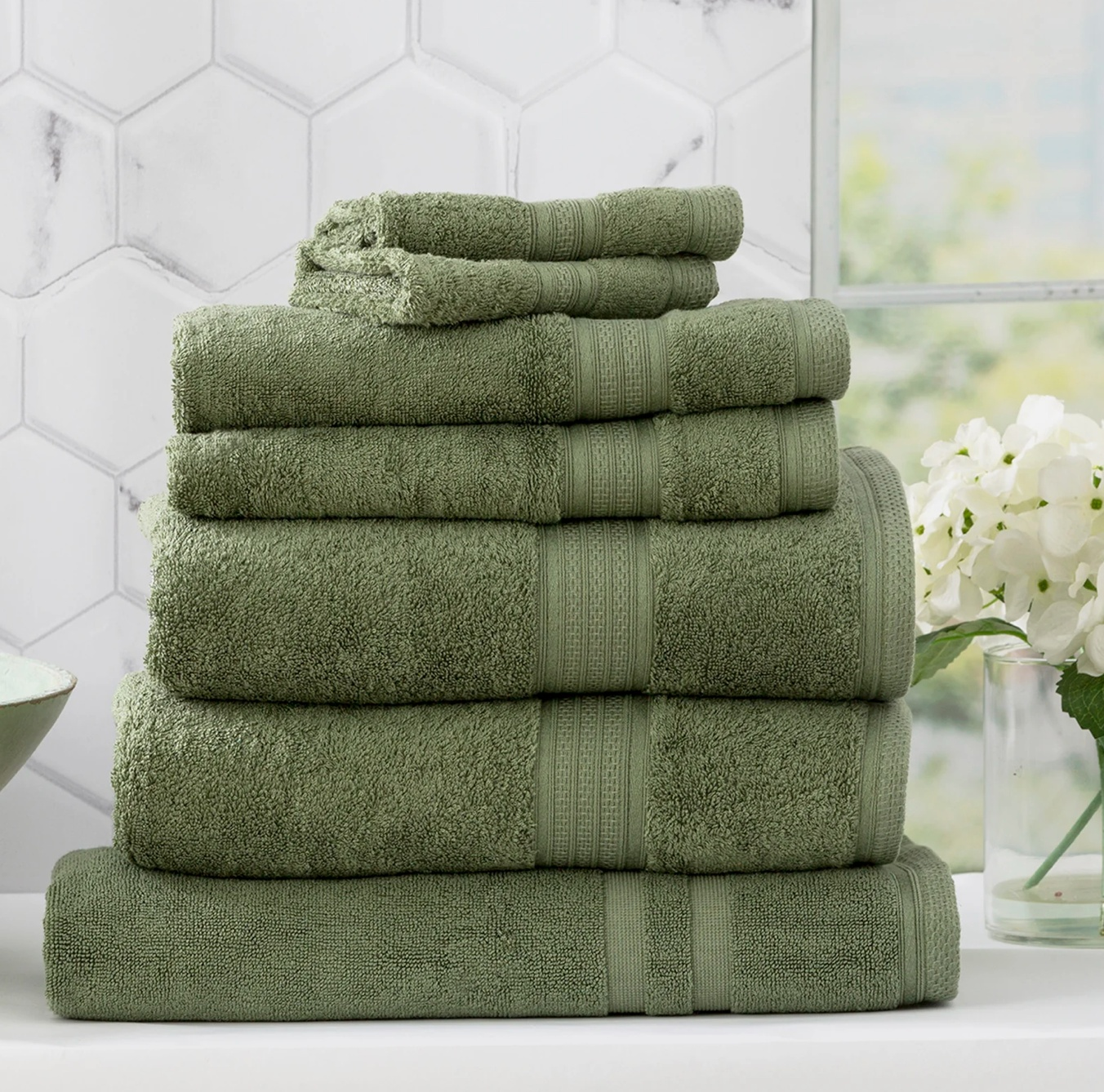 Stack of green towels placed neatly on a bathroom counter with white tiled background and decorative flowers.