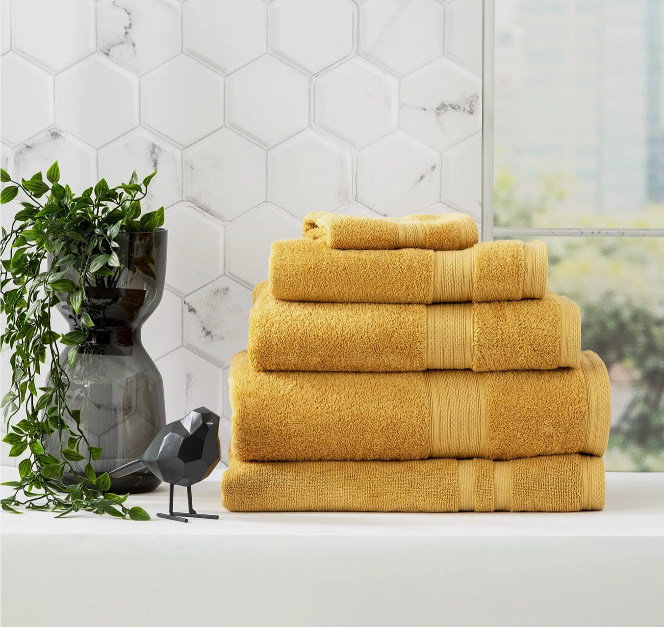 A stack of folded yellow towels next to a black vase with green plant and decorative bird on a white countertop