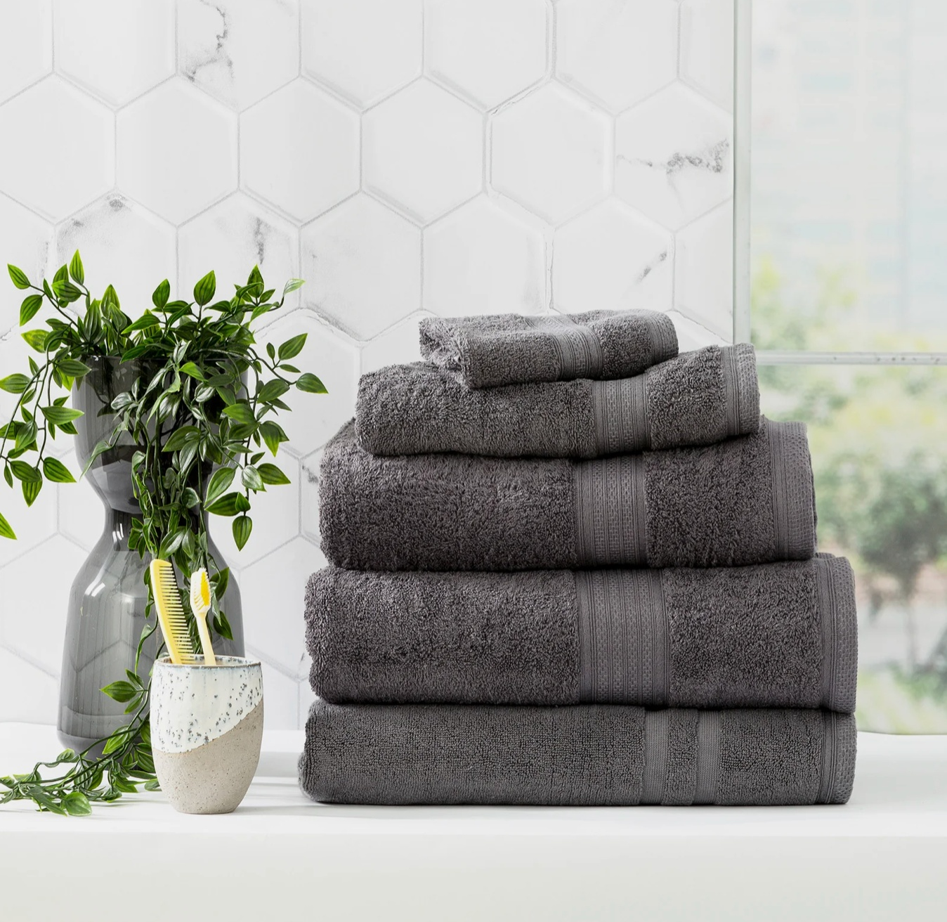 Stack of plush gray towels next to a potted plant and toothbrush holder in a modern bathroom with hexagonal tile backsplash.